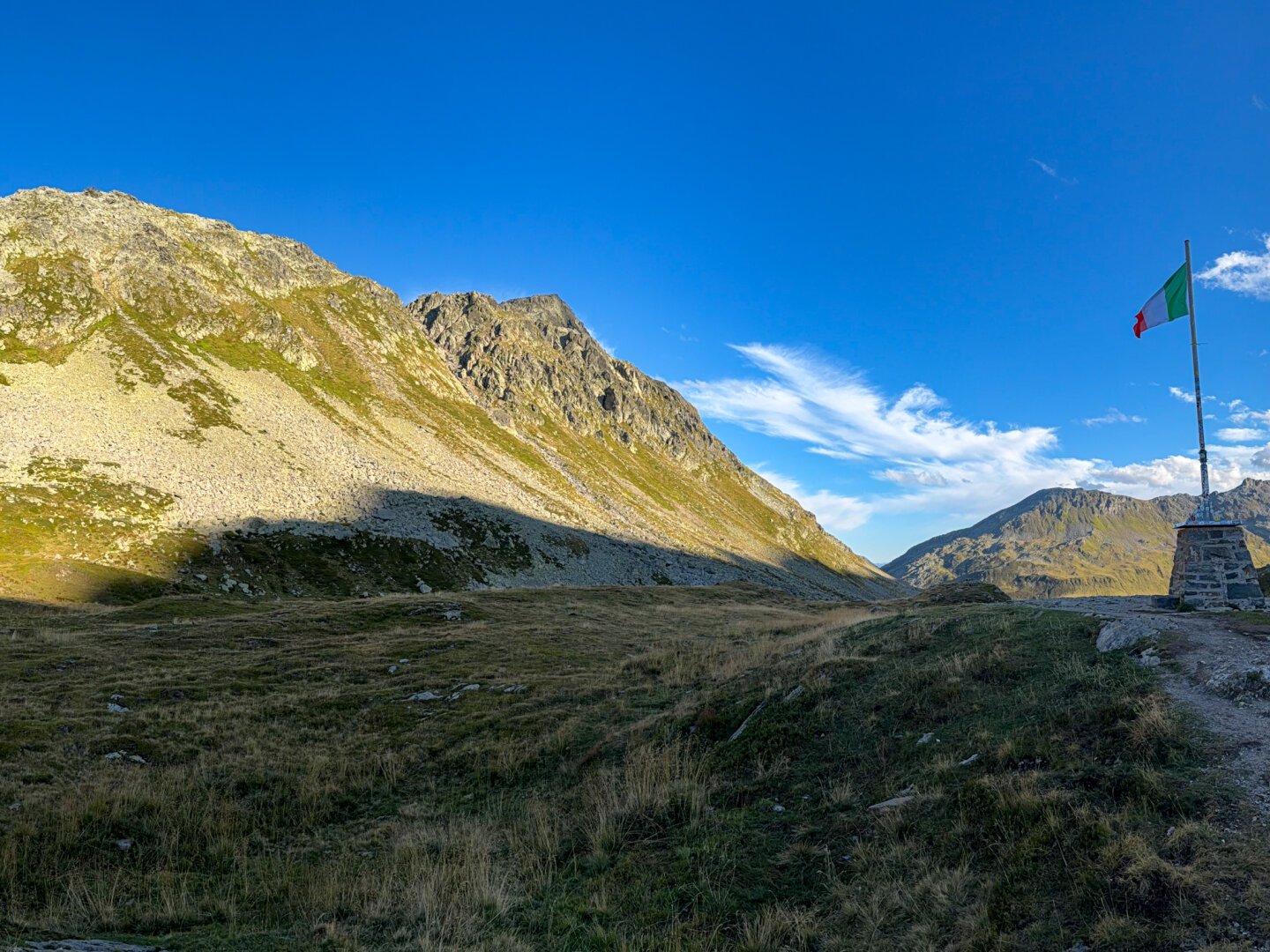 Summit of Passo dello Spluga looking toward Italy. A steep, rocky mountain slope under sunlight, with dark shadows cast over a grassy foreground. The Italian flag flies atop a metal pole set into a stone plinth on the right.