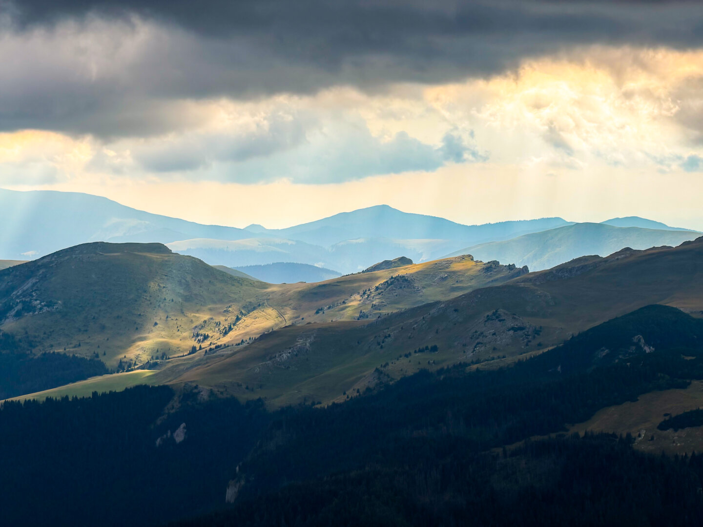 A wide scenic view of layered mountain ridges under a dark, stormy sky. Sunbeams break through a opening in the heavy clouds, casting golden spots of light onto the rolling green slopes and valleys below. Distant blue mountain peaks fade into the soft horizon.