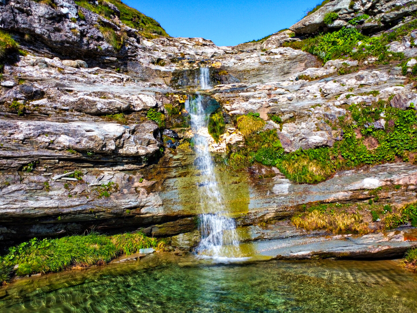 A small waterfall cascades down tiered, rocky cliff ledges into a clear, emerald-green pool below. Lush green moss and mountain grass grow along the cracks in the stone, under a bright blue sky.