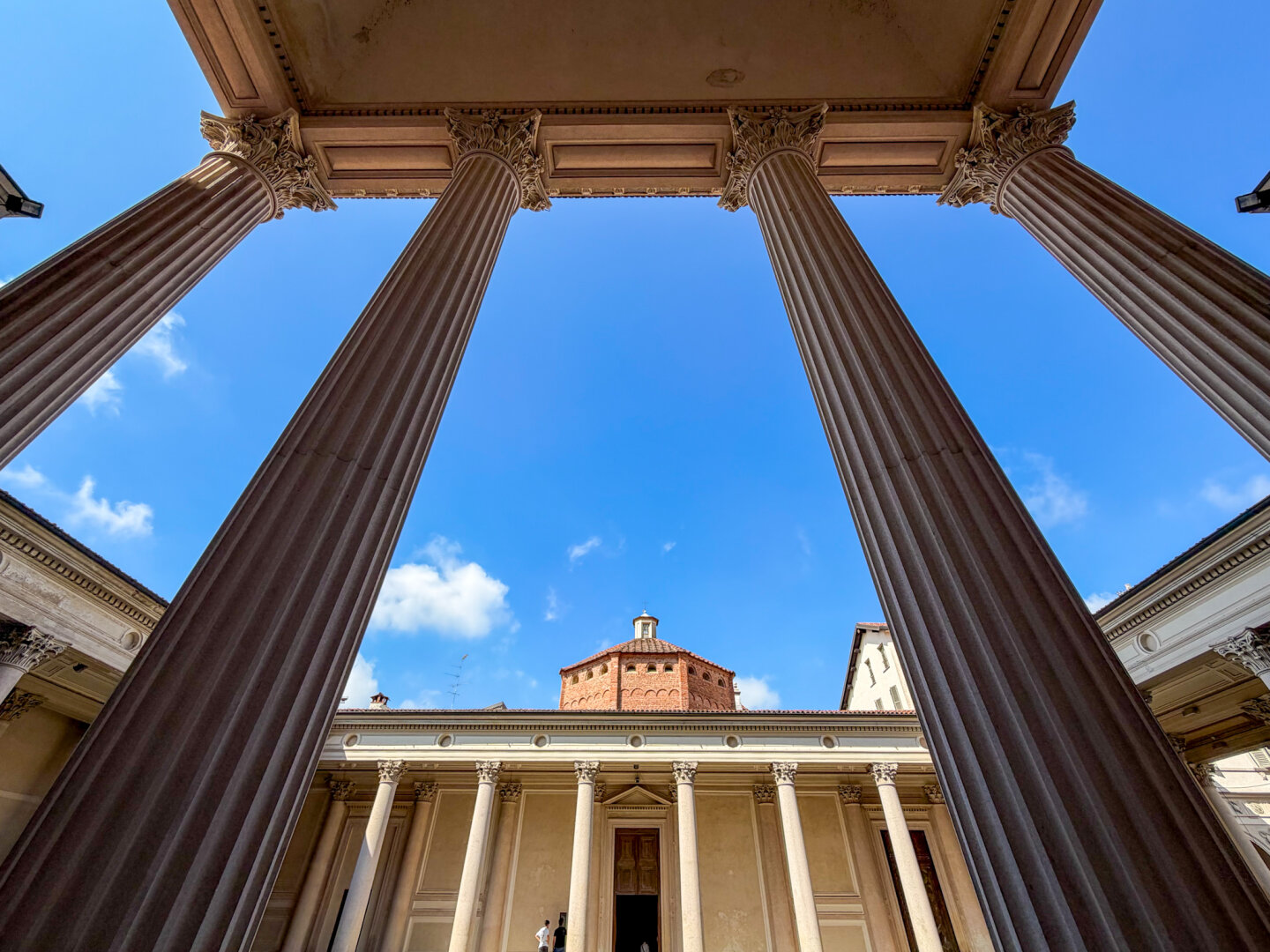A dramatic upward photo taken from beneath a portico, framing tall Corinthian pillars against a vivid blue sky. In the background, a brick dome and surrounding classical colonnades are visible.