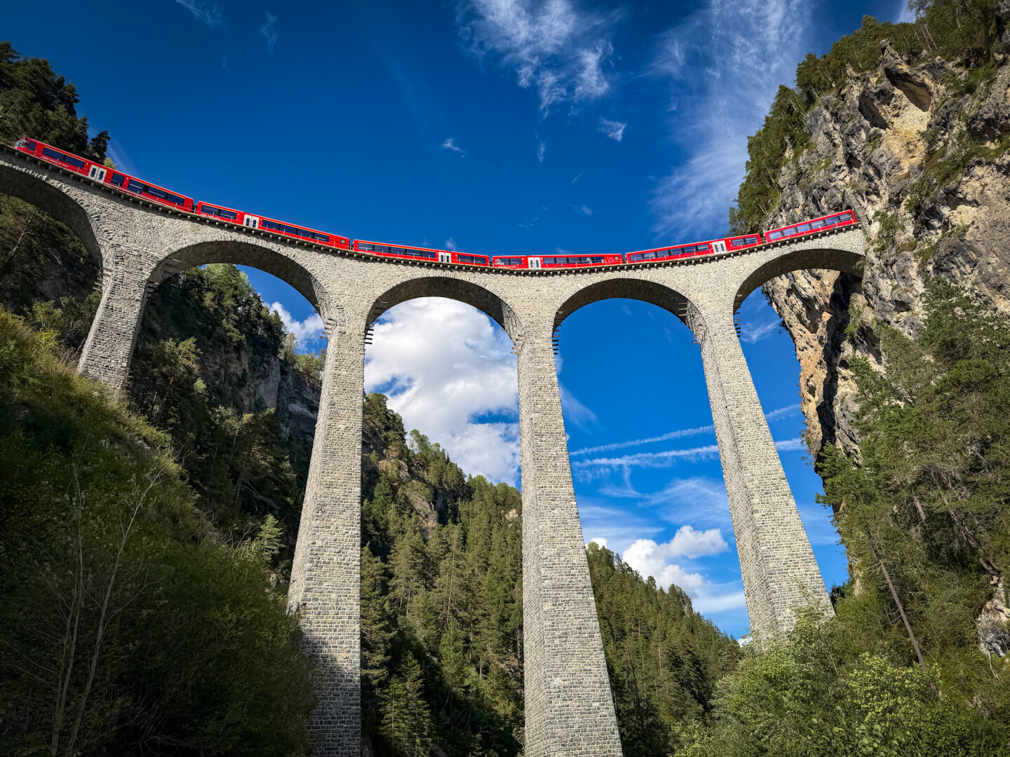 Low-angle shot looking up at the iconic Landwasser Viaduct, a towering stone arch bridge surrounded by steep cliffs and pine trees. A bright red train curves across the top of the bridge under a clear blue sky.