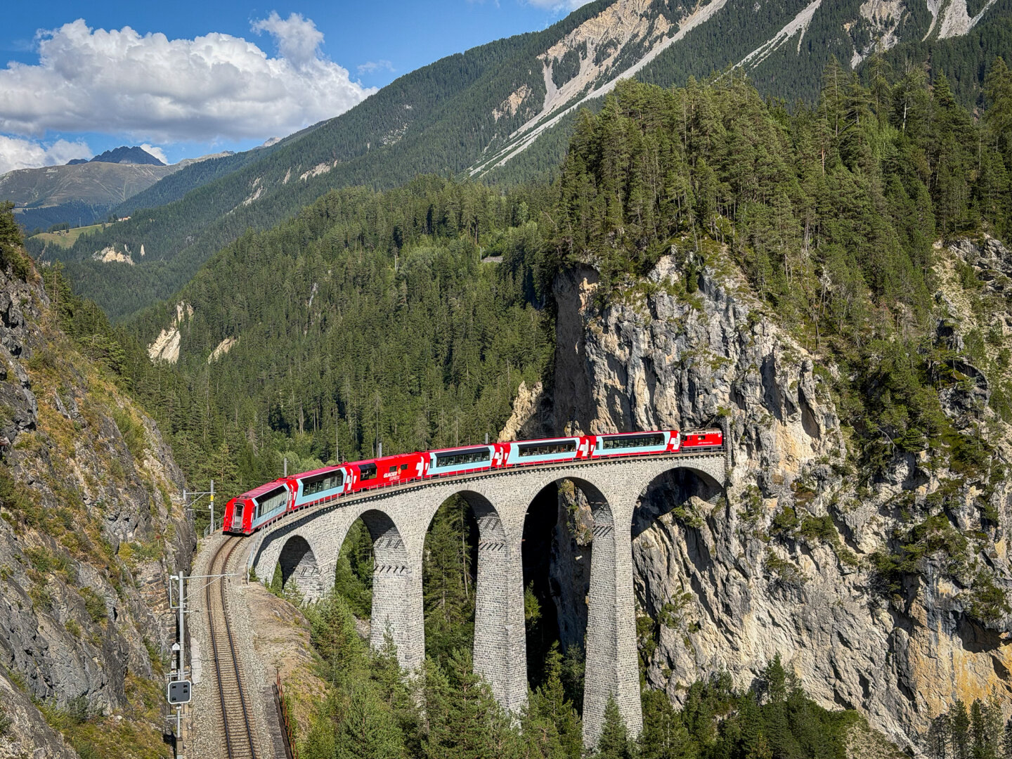 High-angle landscape view of a red Swiss train crossing the curved stone Landwasser Viaduct as it disappears into a tunnel built directly into a steep rock face, surrounded by pine forests and mountain peaks.