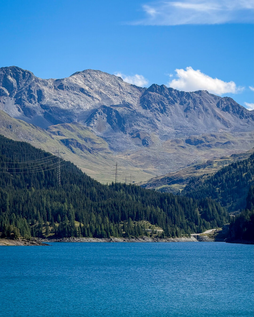 Vertical landscape photo of a calm, deep blue alpine lake framed by a dense dark green pine forest on the shore. Majestic, rocky grey mountain peaks rise steeply in the background under a vibrant blue sky with a few light clouds. Power line towers run along the forested slope leading toward a winding mountain road on the right.