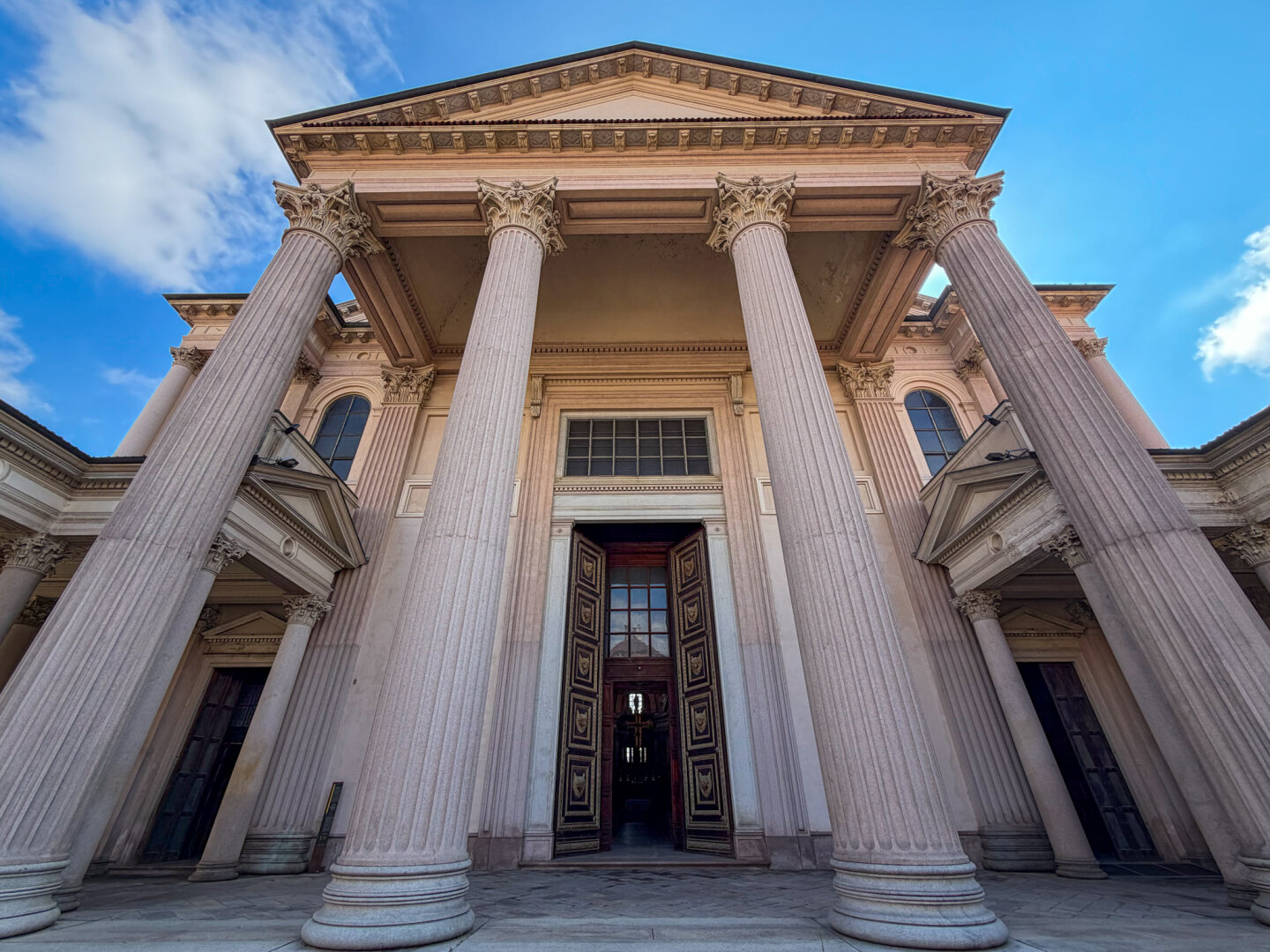 A wide low-angle view of a grand classical building with massive Corinthian columns supporting a triangular pediment. The large wooden entrance doors in the center are open, revealing a dark interior under a partly cloudy blue sky.