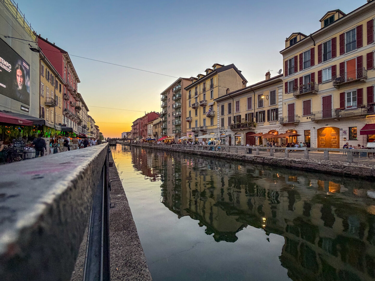 A wide, low-angle shot along a peaceful canal line in Milan's Navigli district during sunset. Pastel-colored, multi-story historical buildings line both banks, reflecting softly in the calm water. Warm sunset light paints the horizon orange and yellow, while outdoor cafes line the walkways, filled with people enjoying the evening.