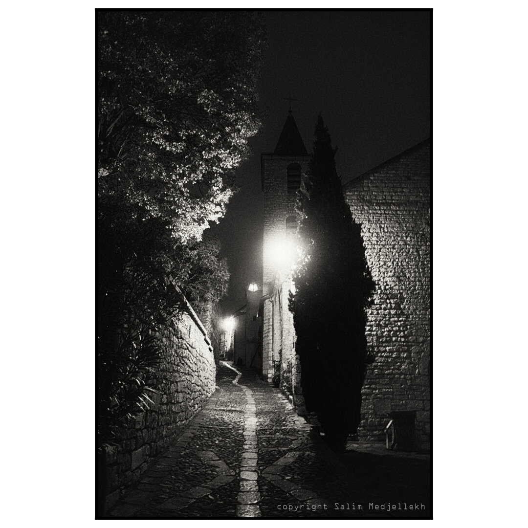 Photographie en noir et blanc de Sainte-Enimie , la nuit,  avec un cyprès éclairé par un lampadaire avec une église en arrière fond.
A black-and-white photograph of Sainte-Enimie at night, showing a cypress tree lit by a street lamp, with a church in the background.