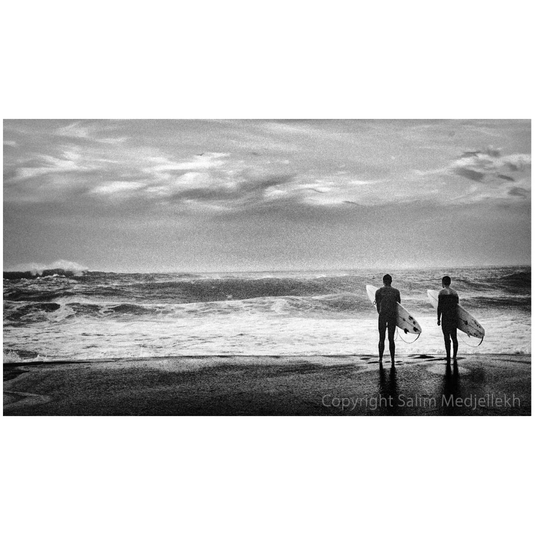 Photographie en noir et blanc de deux surfeurs qui observent l'océan, attendant la vague.
A black-and-white photograph of two surfers gazing out at the ocean, waiting for a wave.