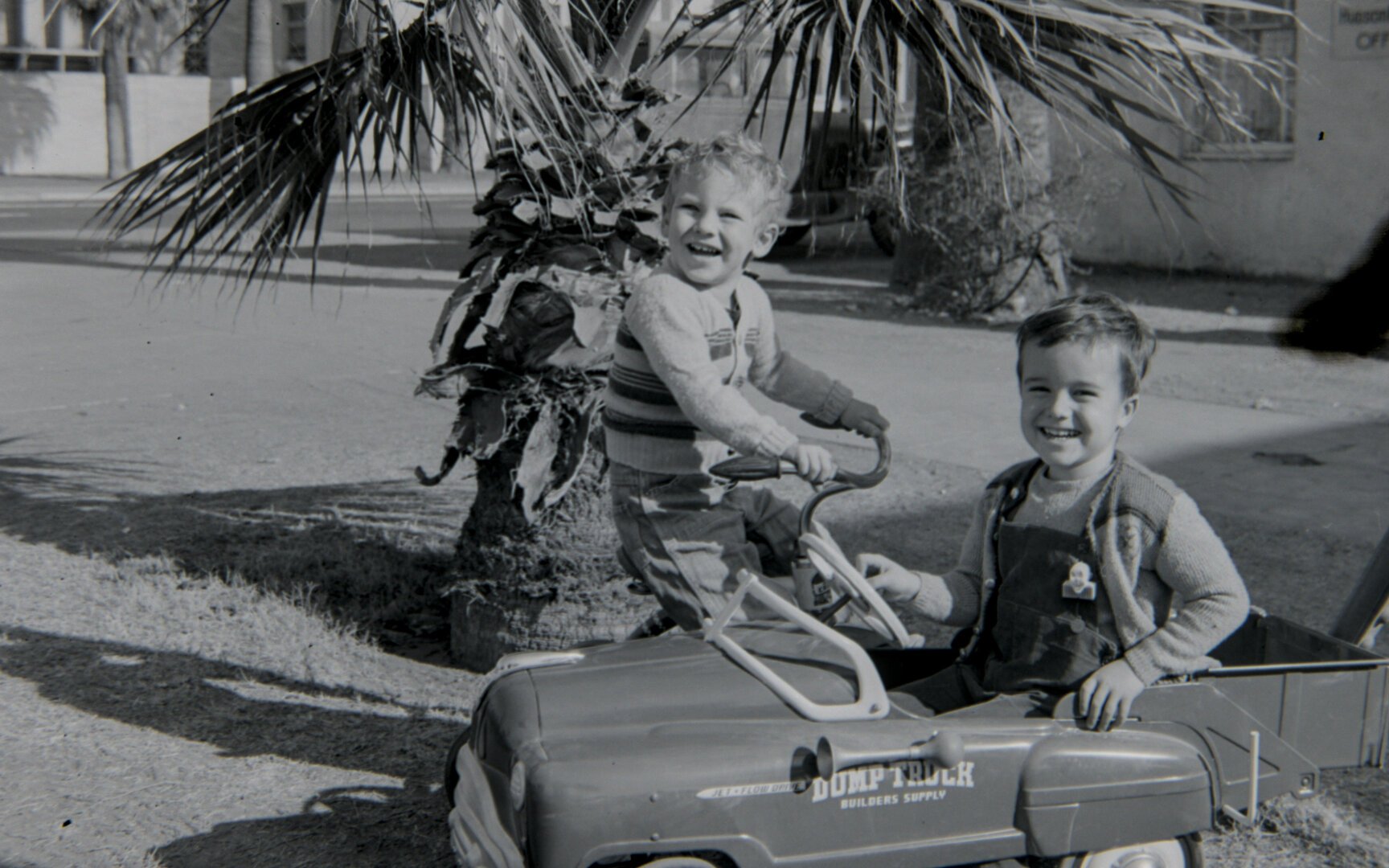 A young boy in a dump truck and a young boy on a tricycle grin at the camera