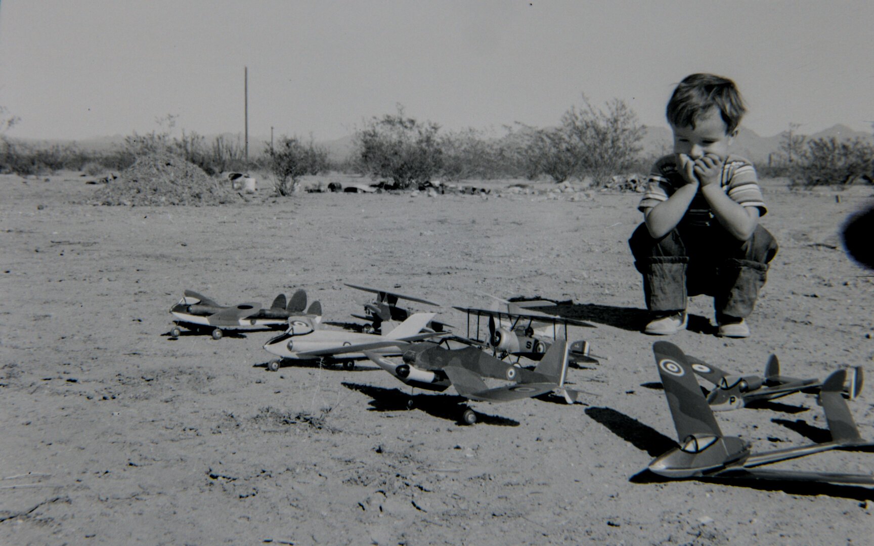 A young boy squatting with his hands on his face, taking in a fleet of model planes laid out in front of him on the ground