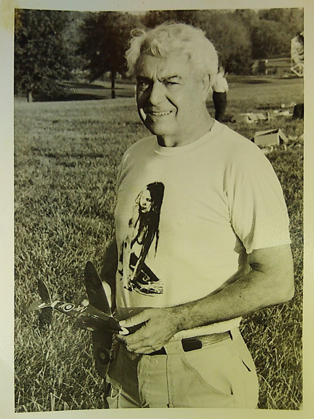 A man with a stylized naked woman on his t-shirt stands in a field, holding a model airplane