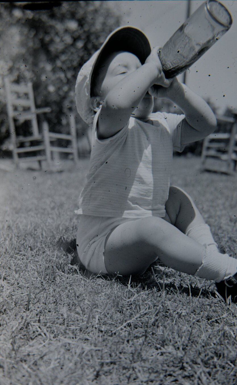 a little boy sits on the ground trying to get the last bit out of a glass bottle