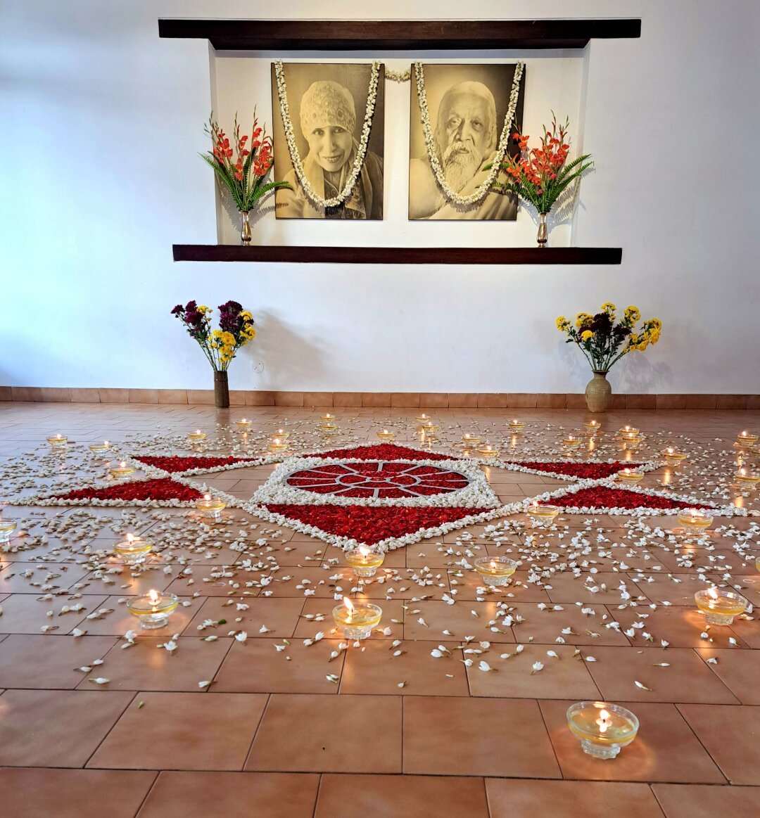 Photography of The Mother and Sri Aurobindo. Surrounded by flower bouquets and arrangement of flowers in the Mother's symbol. Candles surrounding the flower arrangement on the floor.