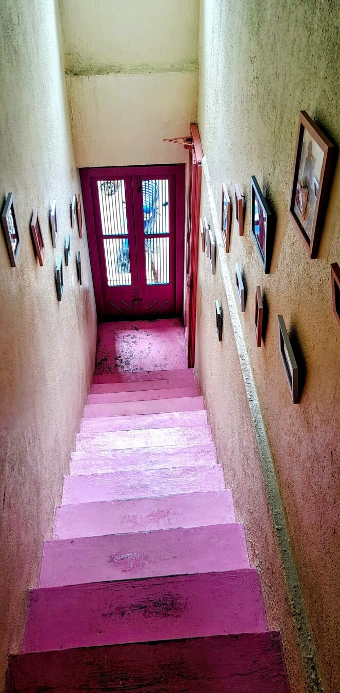 Pink staircase leading to a pink door. Multitudes of Photo frames on both side of walls. Contours of a motorcycle outside the door is visible.