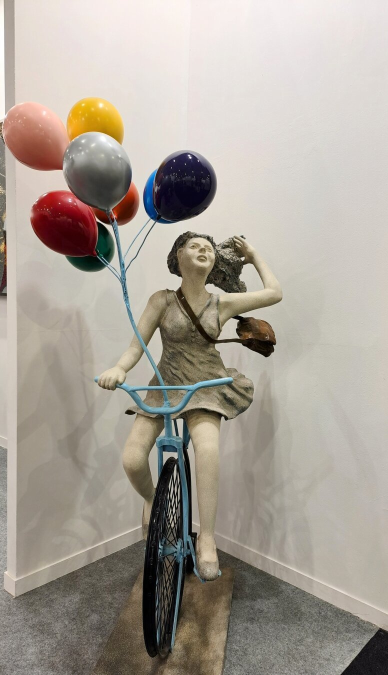 Sculpture of a woman with a satchel on bicycle playing through her hair. 7 multicolored balloons tired to her blue bike.