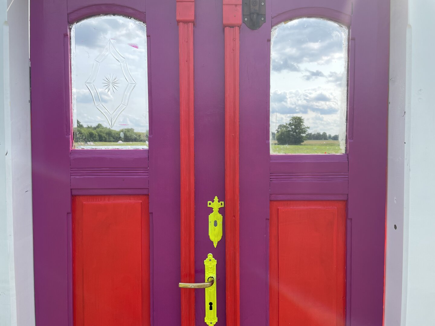 A Front door in purple and red. The door has small windows which Show a landscape. Outside in view

Eine rote und lila Haustür mit Blick von außen. Durch die Fenster ist eine Landschaft zu sehen. Es wirkt so als wäre drinnen draußen.