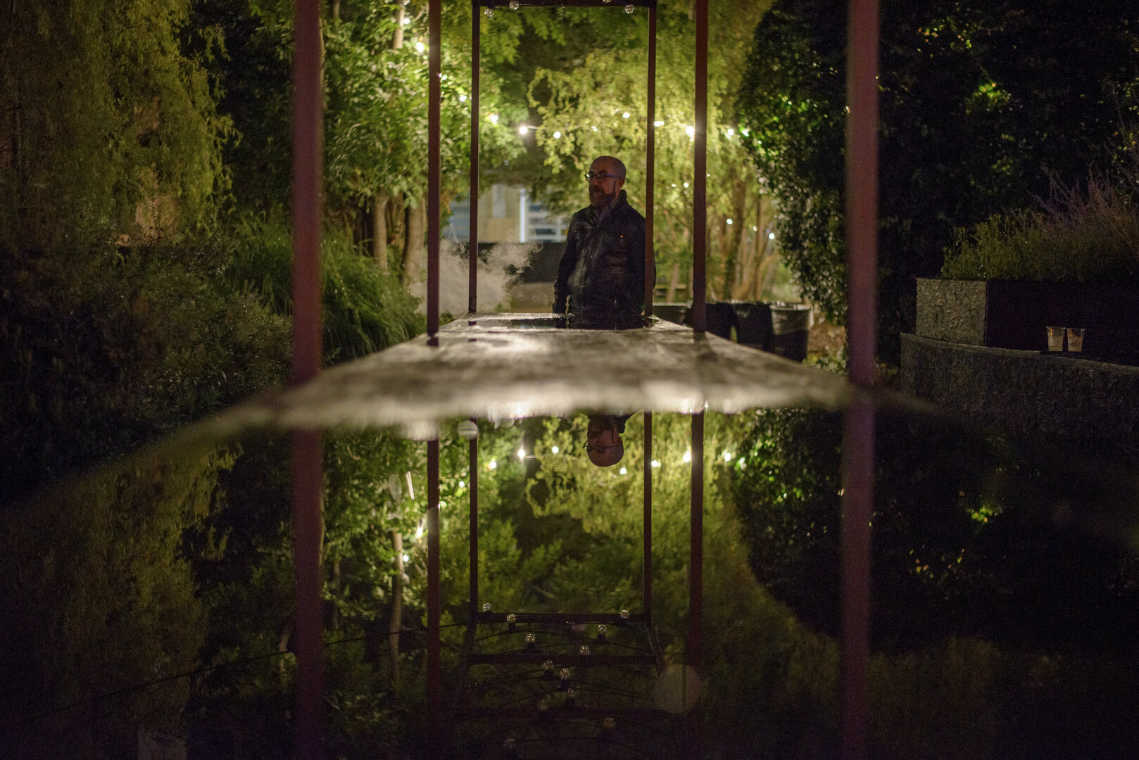 A dimly lit, symmetrical night photograph. A man stands in the background, framed by a glowing rectangular structure. His perfect reflection is visible in the still, dark water of a long rectangular pond that fills the bottom half of the image. Dark, out-of-focus pillars frame the scene in the foreground, creating a sense of depth.