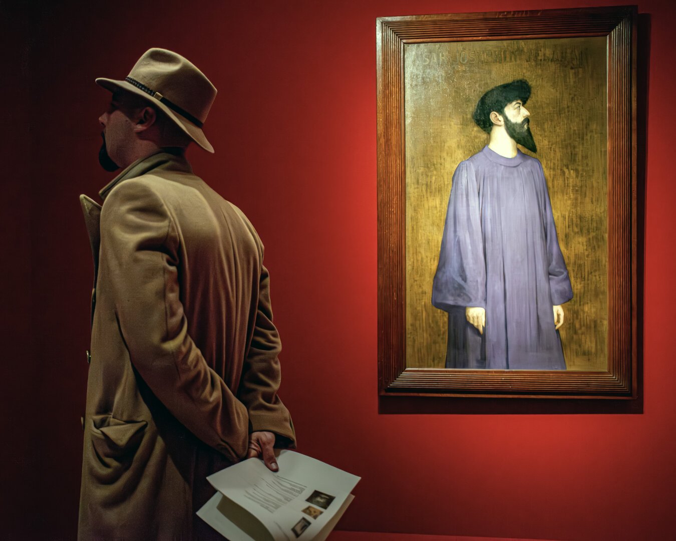 Man in beige coat and fedora views a framed portrait of a bearded figure in a purple robe against a red gallery wall.