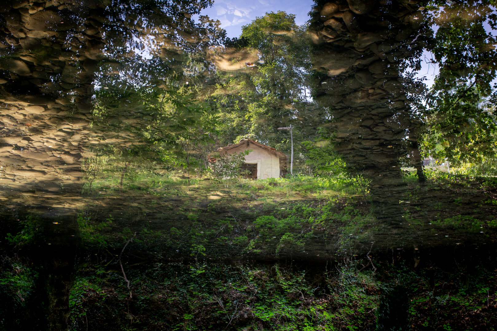 A photograph of a reflection in water. The reflection shows a small, rustic house surrounded by green trees and a patch of blue sky. The riverbed, with visible round stones, is seen through the water at the top and bottom of the frame, creating a layered, painterly effect.