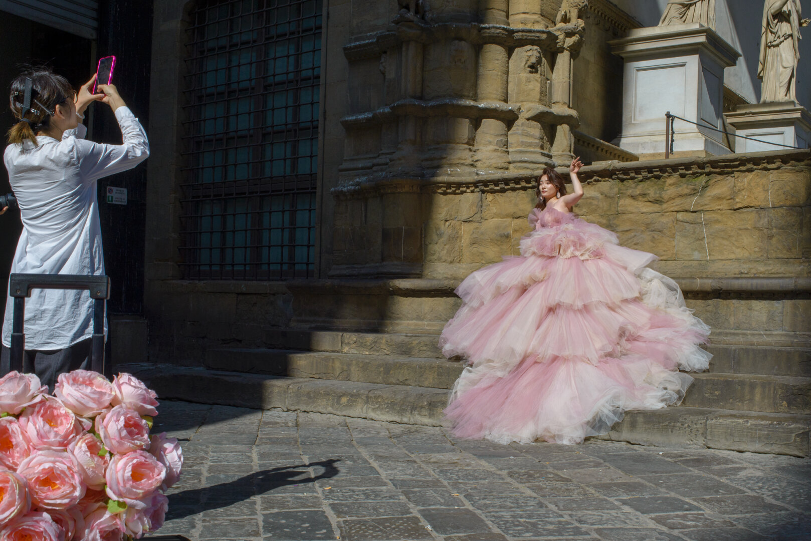 A woman in a large, tiered pink gown poses on stone steps next to an ancient stone building. To her left, another person, seen from behind, holds up a phone, taking a picture. A large bouquet of pink roses is in the lower left corner.