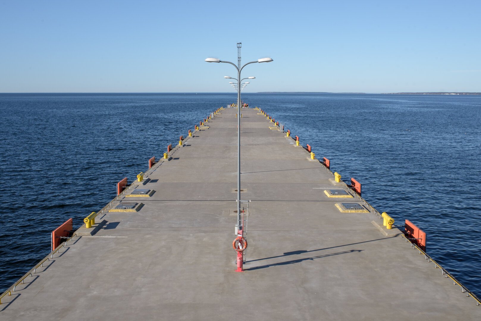 A perfectly symmetrical photograph looking down the center of a long, straight concrete pier extending into a calm, deep blue sea under a clear light blue sky. Identical lamp posts run down the middle, and repeating yellow and orange bollards line the edges. The horizon is flat and distant. Sharp shadows indicate bright sunlight.