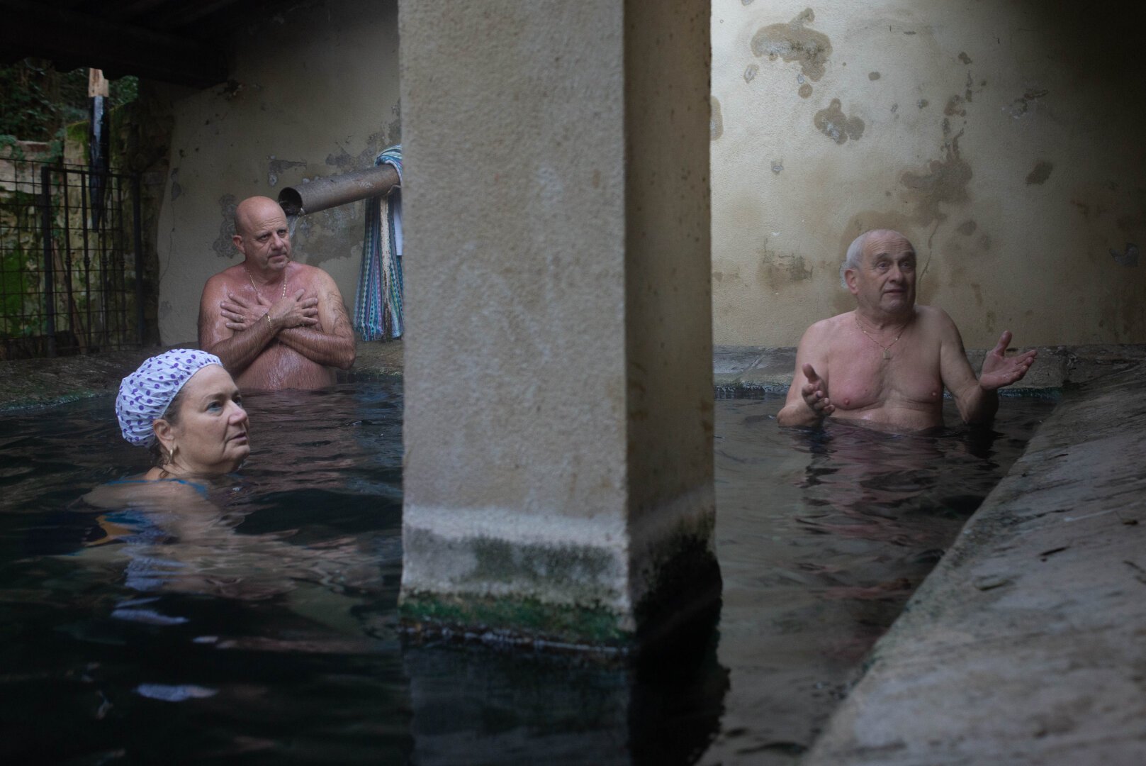 In dim, atmospheric light, three people—a woman in a swim cap looking left, a man behind her, and another man gesturing to the right—are partially submerged in dark water within a rustic stone structure featuring a large central pillar dividing the scene.