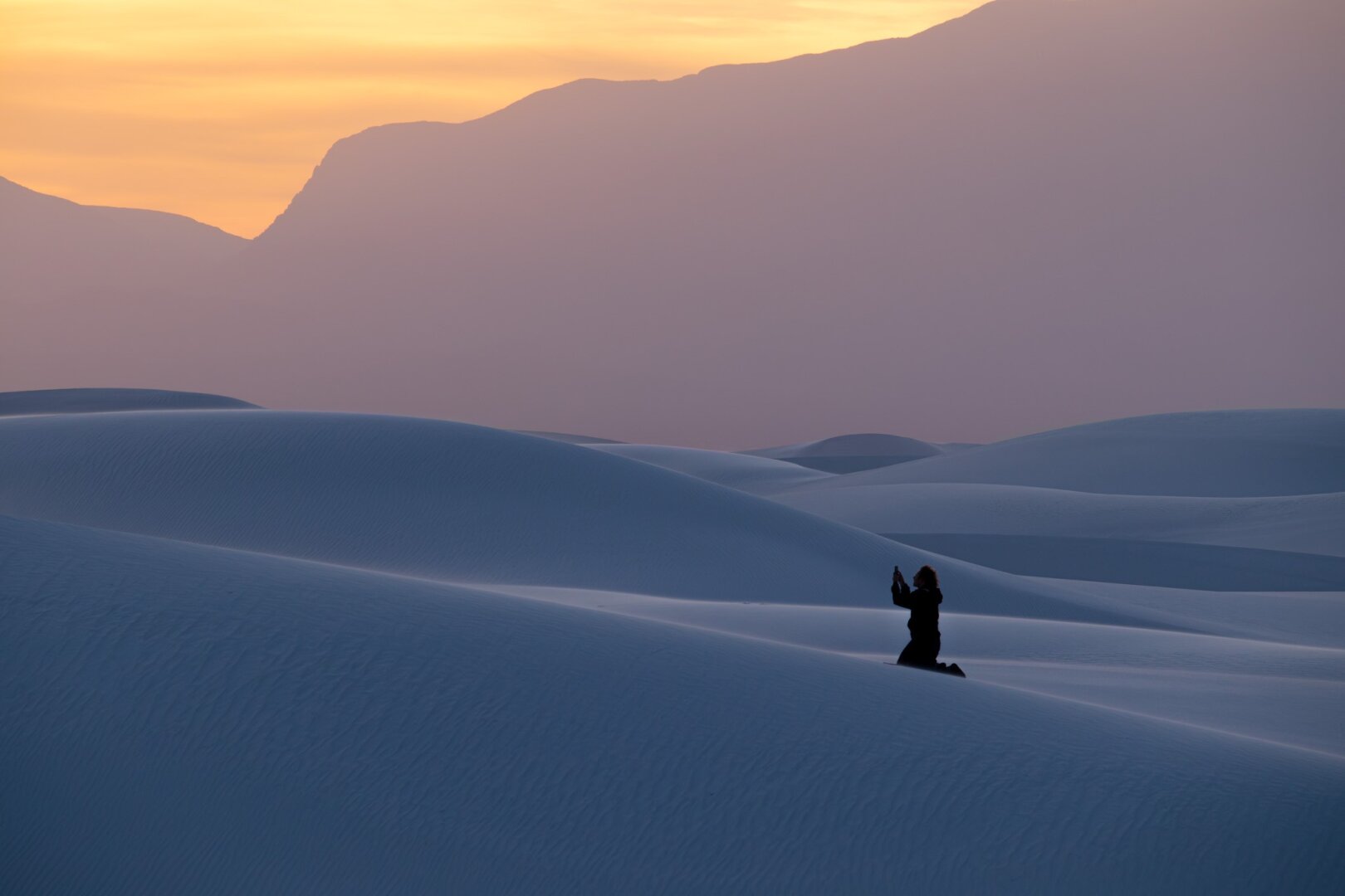 A man on top of the dunes at White Sands, New Mexico sits on his knees for a selfie at sunset.