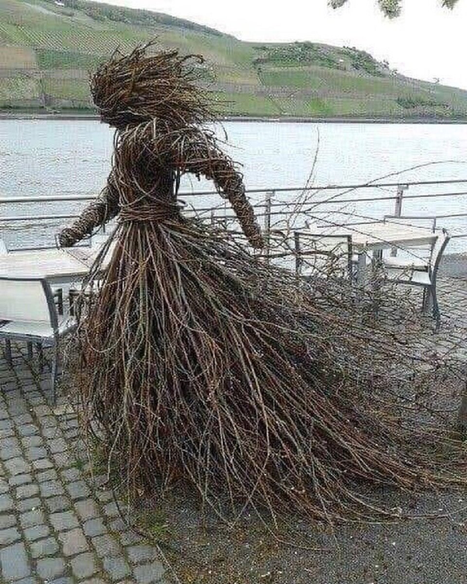 Sculpture of a human figure formed from intertwined branches, standing on a stone terrace beside a lake with green hills in the background.