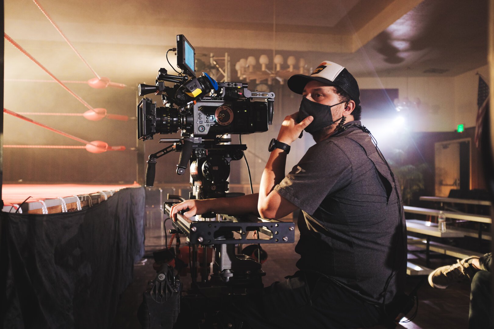 A camera operator looks off camera in preparation for framing his next shot. A cinema camera sits in a slider which is attached to a dolly. On the left is a wrestling ring