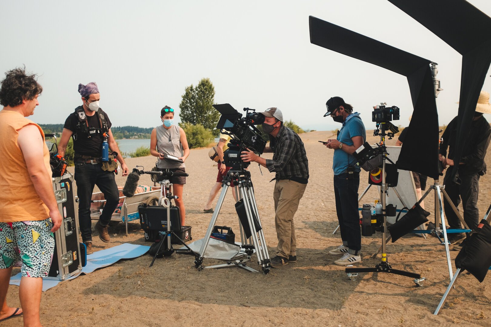 A wide shot of a film crew, cinematographer in center frame. They are on a beach on a sunny day. The cinematographer is framing up his shot