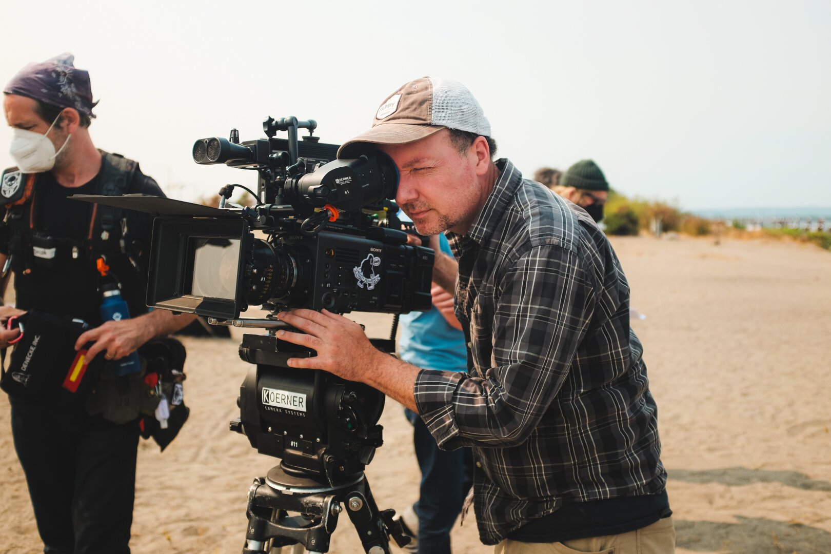 A cinematographer looks in his viewfinder to frame a shot. Behind him are some additional crew. It’s a sunny day in a beach