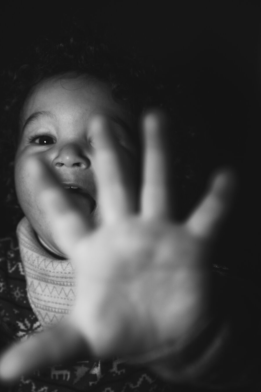 A young child’s hand takes up most of the frame in the foreground and is out of focus, the child’s face and smile, just behind, is in focus. The index and middle finger obscures some of the face. Picture is black and white. No visible background can be seen. Just darkness surrounds a child lit by a flash