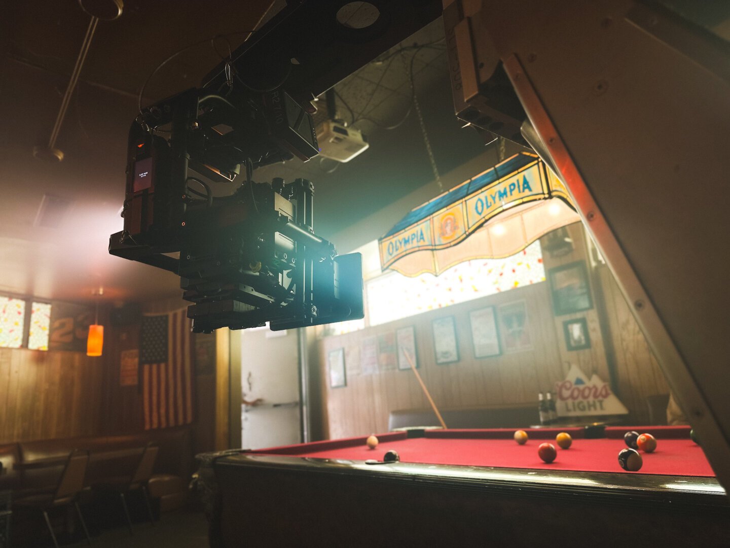 A cinema camera in the foreground hangs underslung on a dolly arm. It is framing up a pool table under a very bright window.