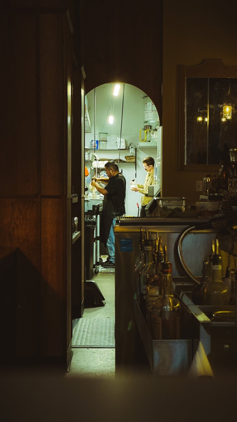 2 men framed through a doorway - stand in a brightly lit kitchen, preparing an order. Outside the doorway it is dimly lit. Bar bottles sit in the lower foreground. The lights in the kitchen give off a green hue from fluorescent lights