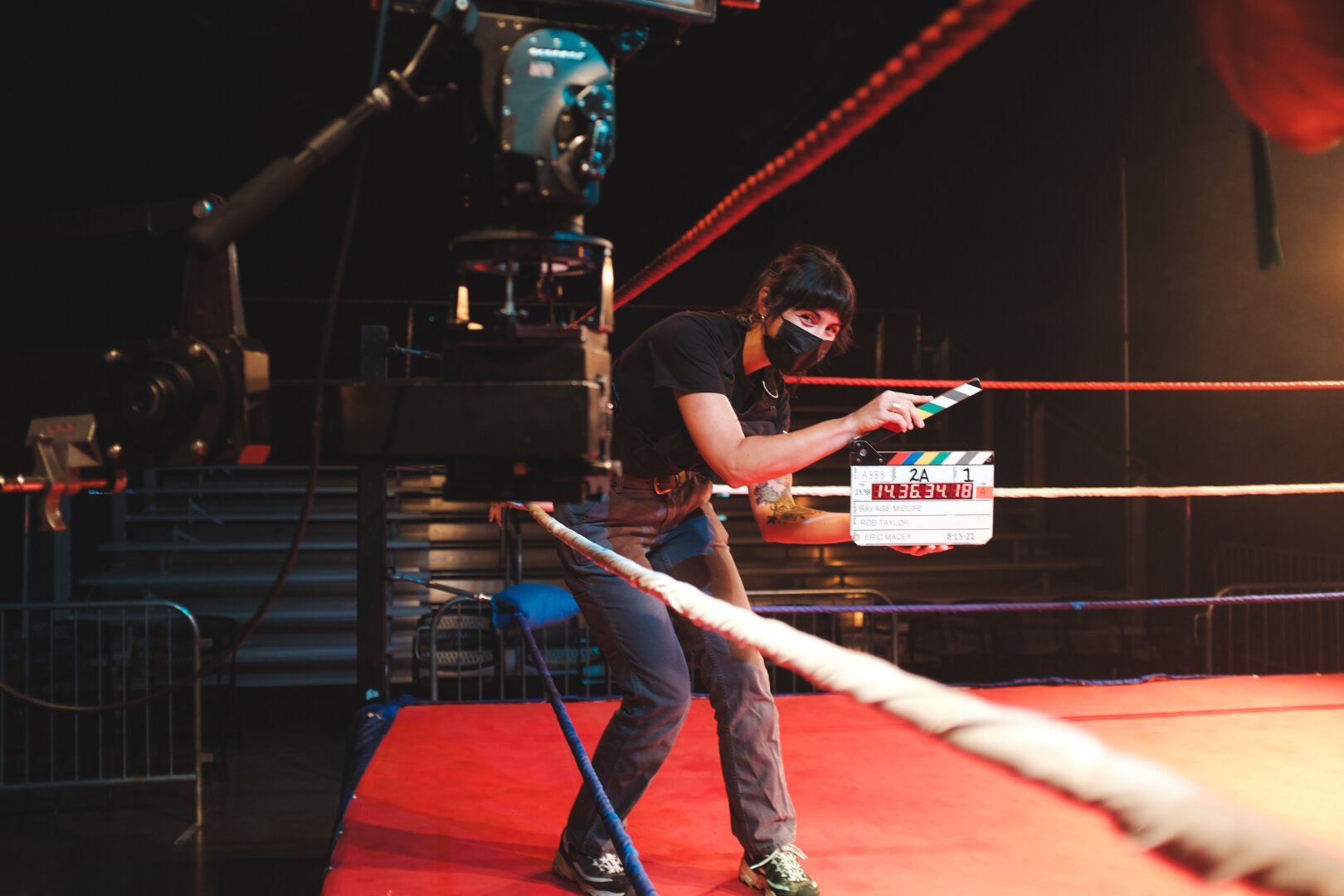 A woman stands in a wrestling ring holding a production slate, ready to clap it for the cameras to roll