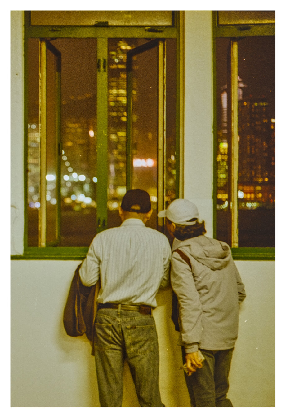 Color photograph taken at the harbor in Kowloon overlooking the bay. Two people, photographed from behind, are standing close together by an open window, looking out at the city at night. One person is wearing a striped shirt and a dark cap; the other is wearing a light-colored hooded jacket and a light-colored cap. Through the tall windows, blurred lights and the illuminated skyscrapers of Hong Kong Island are visible. The grainy photograph is bathed in warm shades of yellow and green.