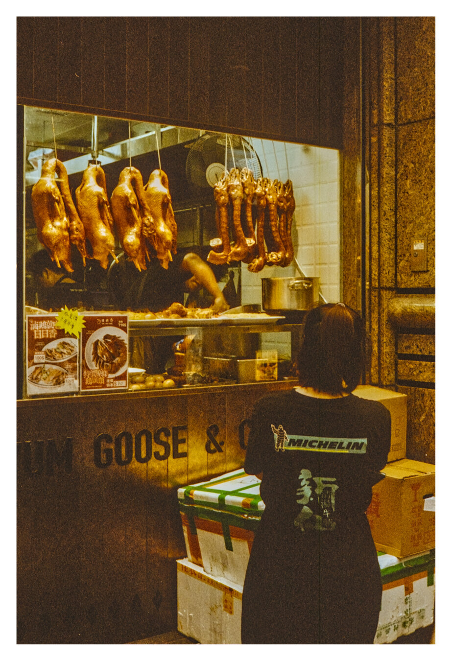 Color photograph of a restaurant display in Hong Kong. In the foreground of the portrait-oriented photo, at the bottom right, a person is standing with their back to the camera, looking at the restaurant window. They are wearing a T-shirt with the word "Michelin" on it. White and brown cardboard boxes are on the floor. Through the window, two people can be seen working in the kitchen. Hong Kong geese (Siu Ngoh) are hanging in the display.