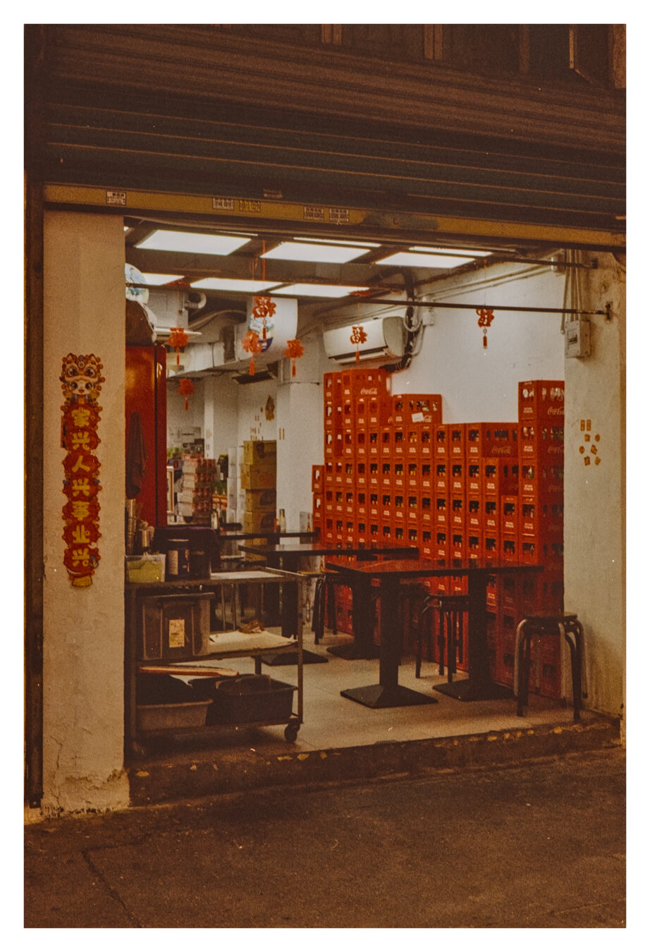 A color photograph taken through an open roll-up door into a simply furnished (street food) restaurant. Numerous red Coca-Cola crates are stacked against the right-hand wall; dark tables and metal stools stand in front of them. Bright ceiling lights and several red Chinese ornaments decorate the room. Hanging from the left-hand entrance pillar is an elongated red-and-gold decoration featuring a lion’s head and Chinese characters that symbolize a traditional Chinese blessing.