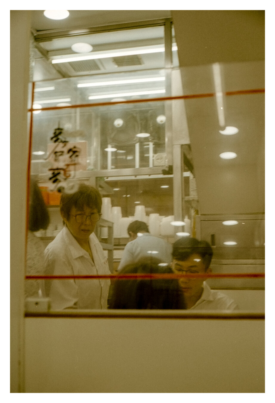 Color photograph of a scene inside a restaurant in Kowloon, Hong Kong. The view looks through a glass pane with red horizontal lines toward a brightly lit kitchen. Behind the reflective glass, two people are seen sitting, and a waitress is standing by the table. Further in the background, parts of the kitchen are visible, including a man working and white plastic containers stacked on the shelves. The photograph is rendered in warm, yellowish tones.