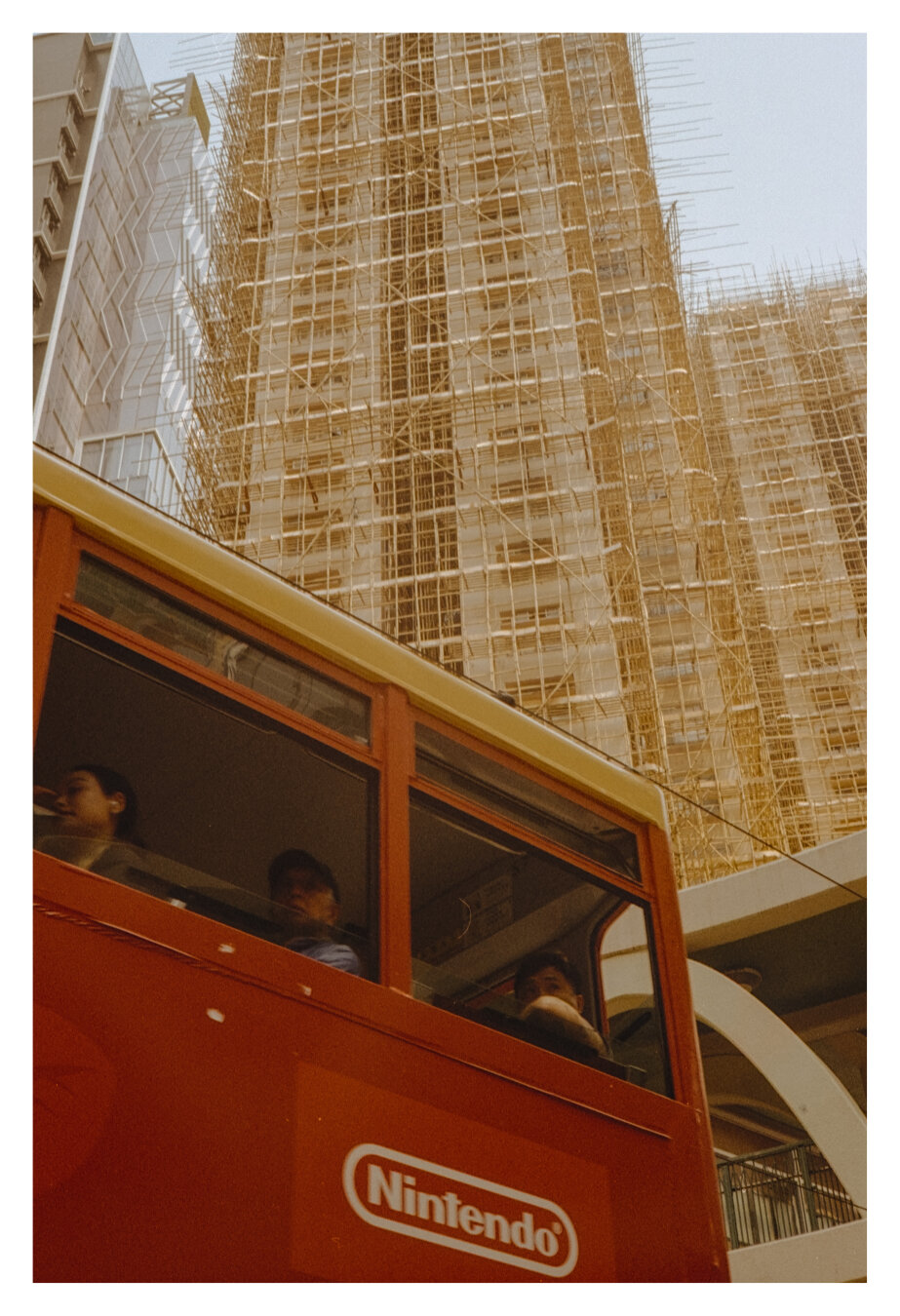 Color photograph of a Hong Kong skyscraper with part of a double-decker tram. The upper half of the portrait-oriented image shows the upper deck of a red double-decker tram. The windows are open, and three people are visible. Below the window, the word "Nintendo" is legible. Behind the tram rises a high-rise building, which is covered in scaffolding in the bamboo-style construction typical of Hong Kong.