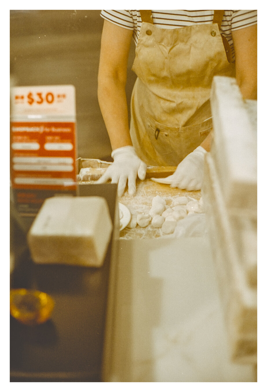 Color photograph taken at a Hong Kong restaurant. The image shows a close-up of a person wearing a striped T-shirt, a light-colored apron, and white gloves, rolling out dough. Several small pieces of dough lie on the floured work surface, which have been shaped into delicious dumplings. Blurred elements of the dining area and kitchen frame the warm, yellowish-toned photograph.