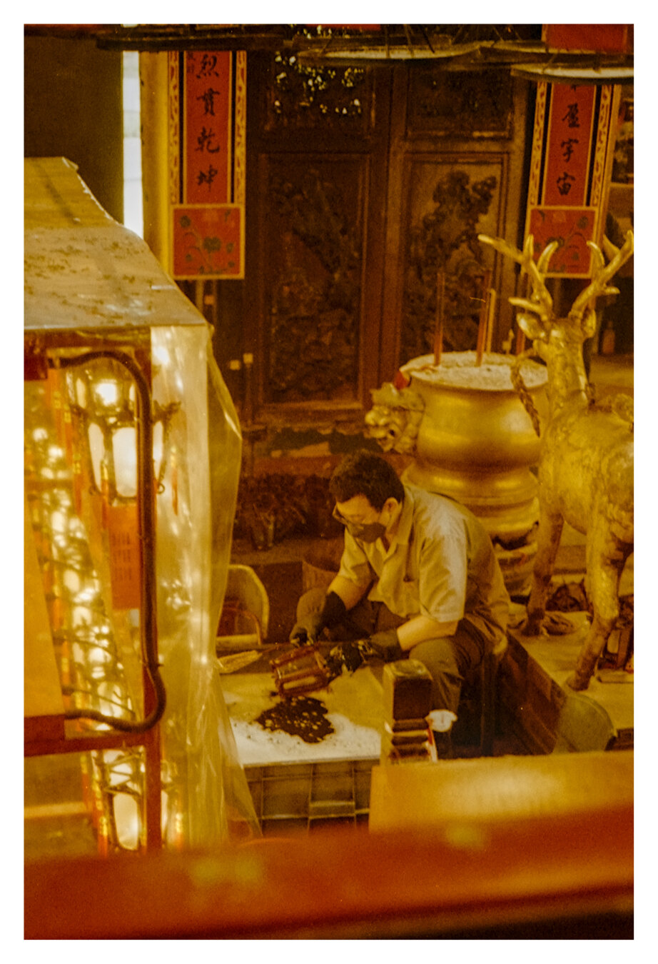 Color photograph taken inside a Hong Kong temple. A person wearing glasses and work gloves is sitting at a low work surface inside the temple, cleaning candle shades with a tool. In the background, a large bronze-colored incense burner and a golden deer figurine stand in front of dark carved wooden doors adorned with red Chinese calligraphy banners. A brightly lit glass display case occupies the left edge of the frame. The photograph is bathed in warm, golden-yellow light.