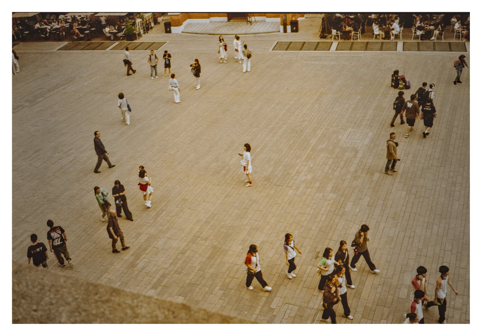 Color photograph of the Parade Ground at Tai Kwun in Hong Kong. It shows a bird's-eye view of the expansive, light-colored paved plaza at the former police station. Numerous people are walking across the open space, either alone or in small groups, while at the top of the frame, others are sitting on benches and steps. In the center of the square—and of the image—stands a person, seemingly completely still, while everyone around them is in motion.