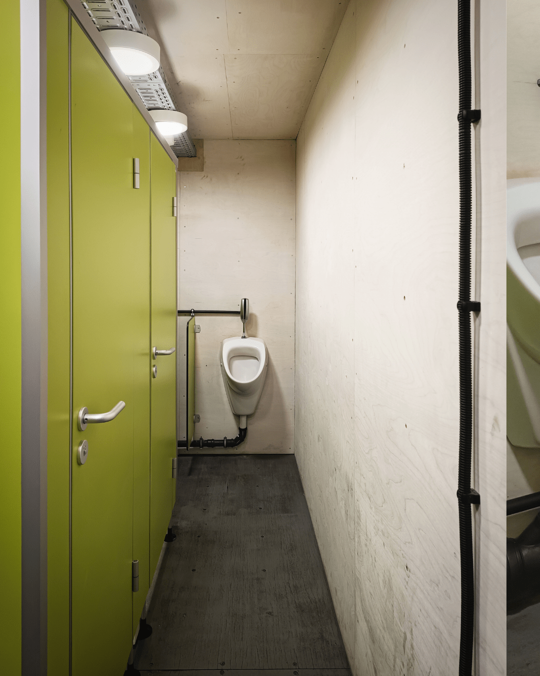 A narrow public restroom with three light green stall doors aligned on the left side. At the end of the corridor, a white ceramic urinal is mounted on the beige wall. The floor is covered with grey tiles, and the ceiling features three recessed lights near the top left corner. The overall lighting is bright and even.