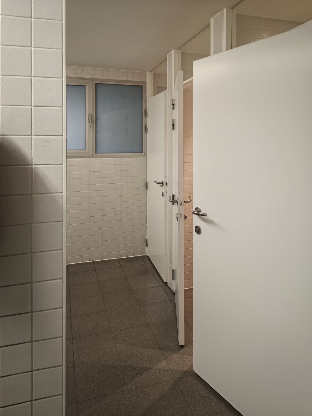 A corner of a public restroom with two white toilet stall doors slightly ajar, revealing tiled interiors. The walls are fully covered in off-white square tiles, and a frosted window allows diffuse daylight into the otherwise dimly lit space. The floor is dark grey tile, and the overall design is minimal and utilitarian.