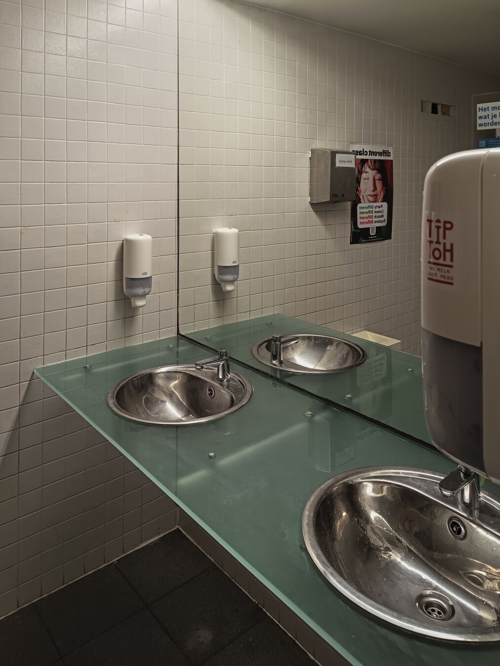 The sink area of a public restroom with a large horizontal mirror mounted above a sleek glass countertop. Two round stainless steel sinks are embedded in the counter, each with a modern single-lever faucet. Liquid soap dispensers are fixed to the tiled wall, and a hand dryer partially obscures the right side. A hygiene awareness poster is visible in the mirror's reflection, along with other restroom fixtures.