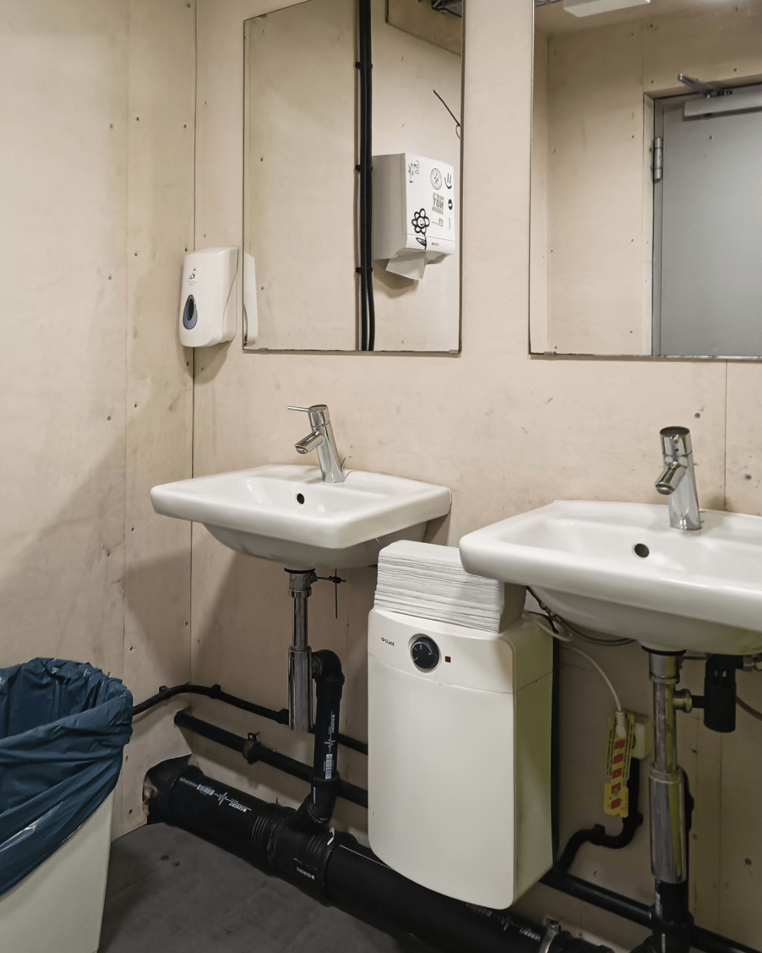 A corner of a public bathroom with two white ceramic sinks mounted against a pale wooden wall. Each sink has a faucet and a mirror above it. Below the sinks, black exposed plumbing and a compact white water heater are visible. A wall-mounted paper towel dispenser and a soap dispenser are also present. A white trash bin with a dark blue liner stands to the left of the sinks. The space appears functional but somewhat industrial in design.