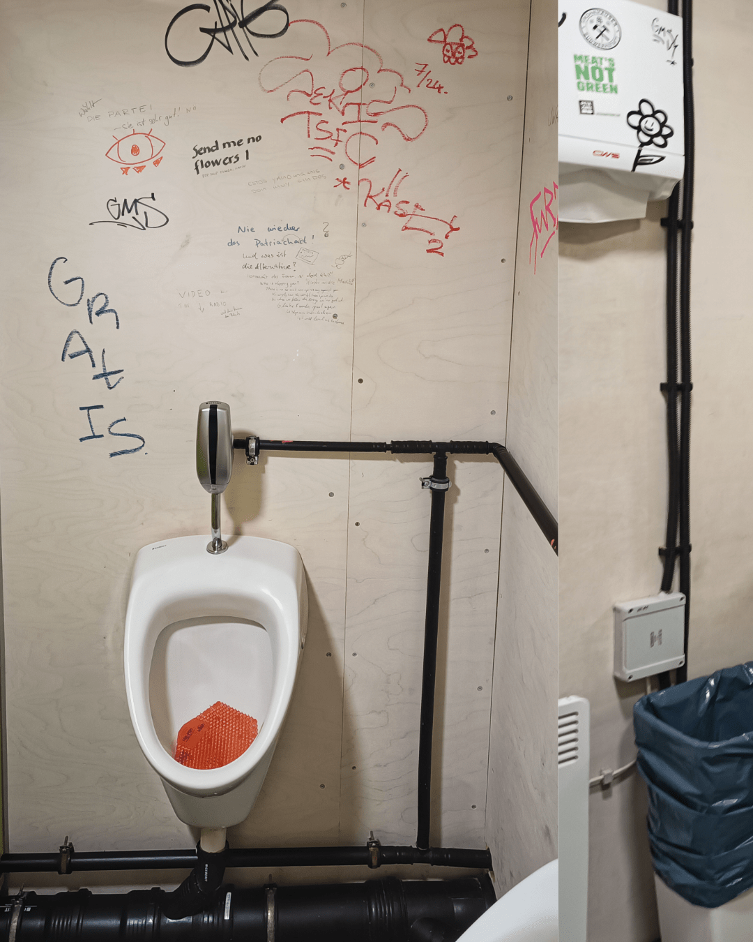 A single white urinal is mounted on a pale wooden wall in a public restroom, with a bright orange urinal mat placed inside it. Above the urinal is a metallic flush valve connected to black piping. The surrounding wall is heavily covered in graffiti and handwritten messages in various colors and styles. To the left, part of a green privacy divider and another urinal are partially visible. The floor and plumbing pipes have a rugged, industrial look.
