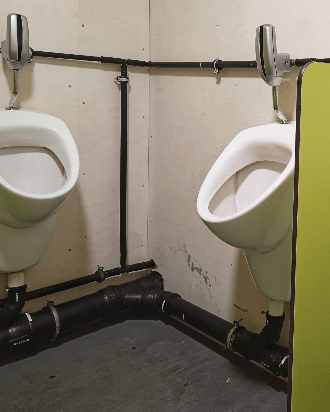 Image part 1 of 2
Three white ceramic urinals are mounted along two intersecting pale wooden walls in a public men's restroom. Each urinal has a metallic flush valve above it, connected by exposed black piping that runs along the wall. A lime green privacy divider separates the middle urinal from the one on the right. The flooring is dark grey, and the overall appearance is utilitarian with visible plumbing fixtures.