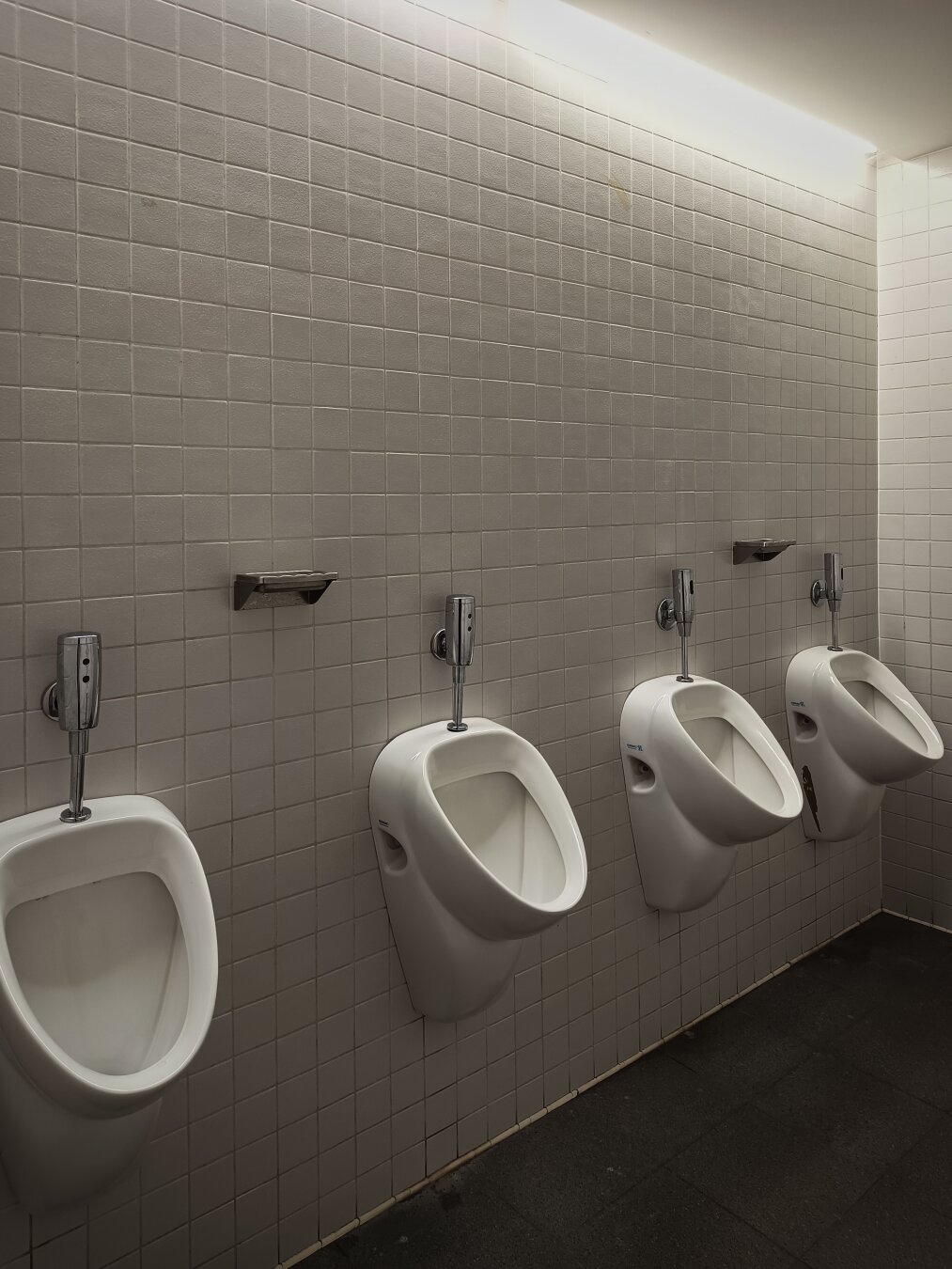 A clean, well-lit public men's restroom featuring a row of four white ceramic urinals evenly spaced along a wall covered with light beige square tiles. Above each urinal is a chrome flush valve, and small metal shelves are mounted between them. The floor is dark grey tile, and the lighting from above casts soft shadows on the wall.
