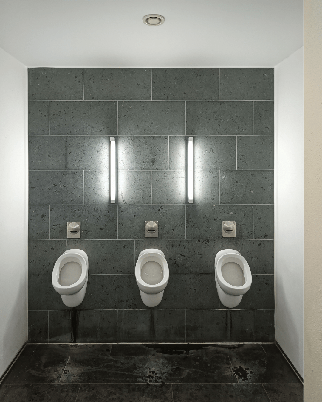 A men's restroom with three white urinals mounted on a dark green tiled wall. Above each urinal is a flush button. Two vertical wall lights illuminate the area. The floor is dark grey with visible water stains.